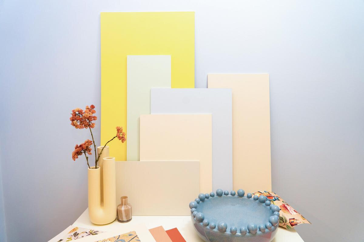Yellow, pink and blue pastel coloured cards propped against a blue wall. A vase of flowers and blue bowl sits in front.